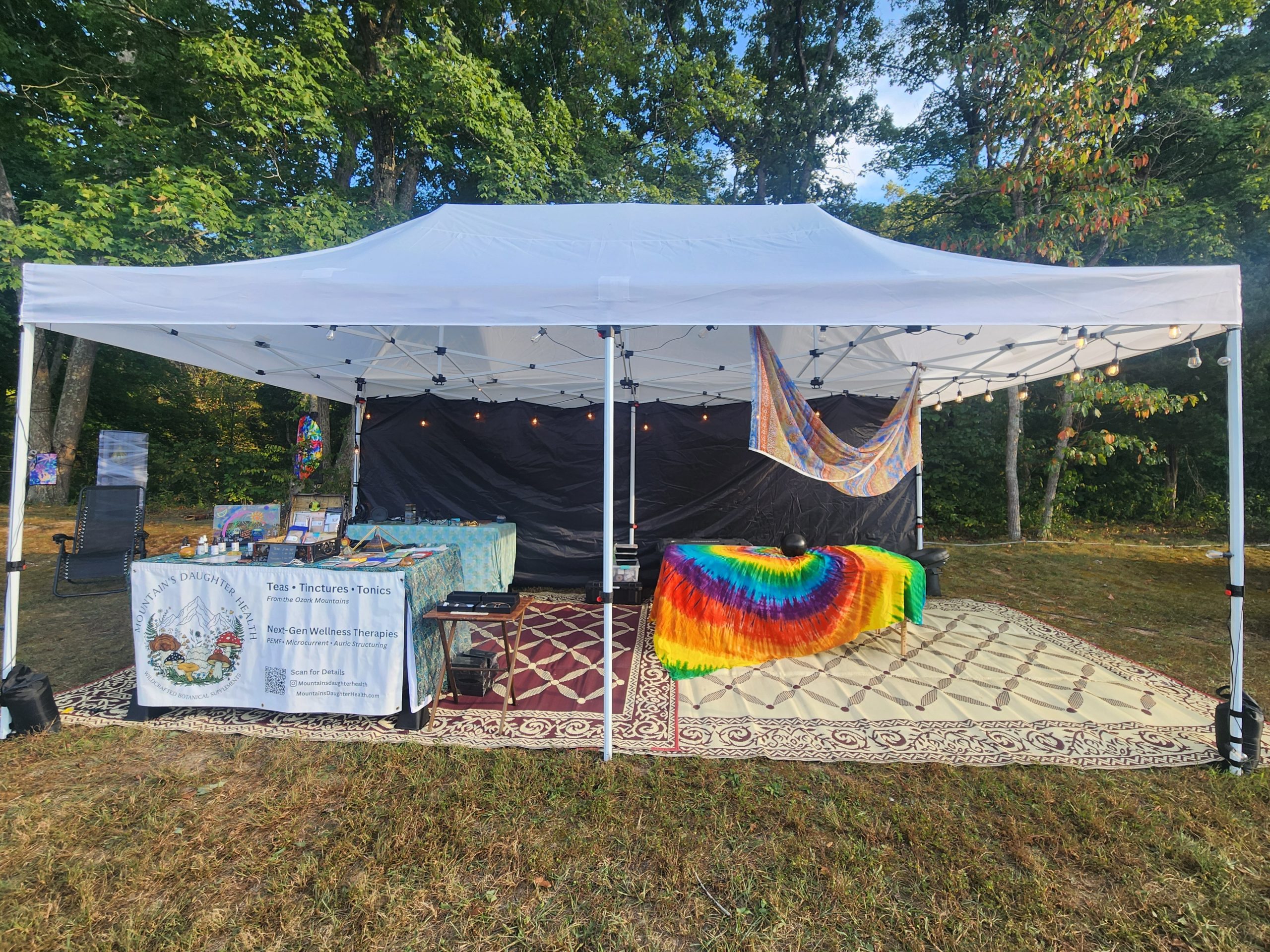 White canopy wellness tent on a lawn with rugs, a tie-dye tapestry, and a Mountain's Daughter Health table under the trees