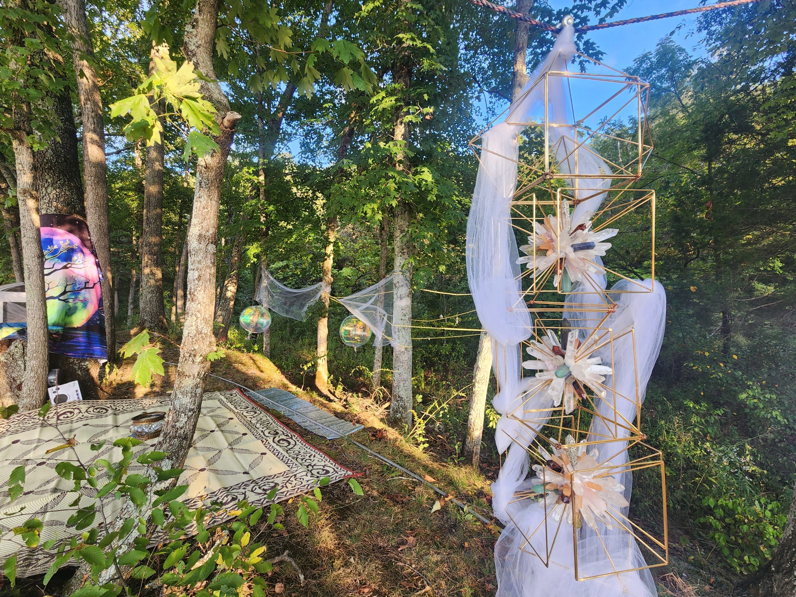 Gold geometric frames holding crystal clusters, wrapped in white fabric among trees in daylight